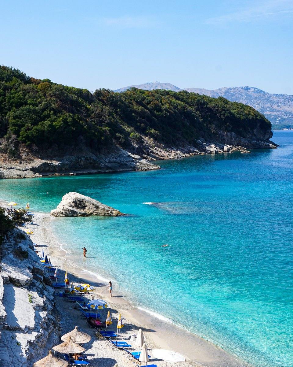 A turquoise cove with a pebble beach and sun loungers below green headlands on the Ionian coast