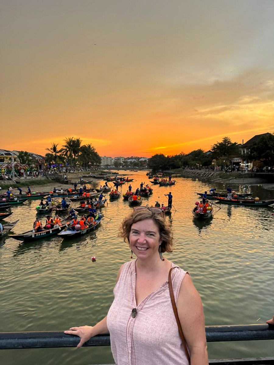 Mairin on a bridge over the Hoi An river at sunset, with sampan boats drifting on the orange water