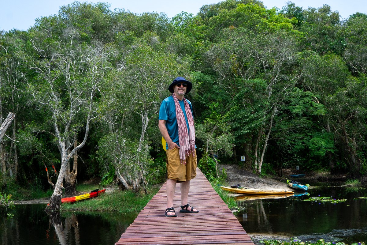 Todd standing on a wooden jetty over a still forest lake with kayaks pulled up on the bank